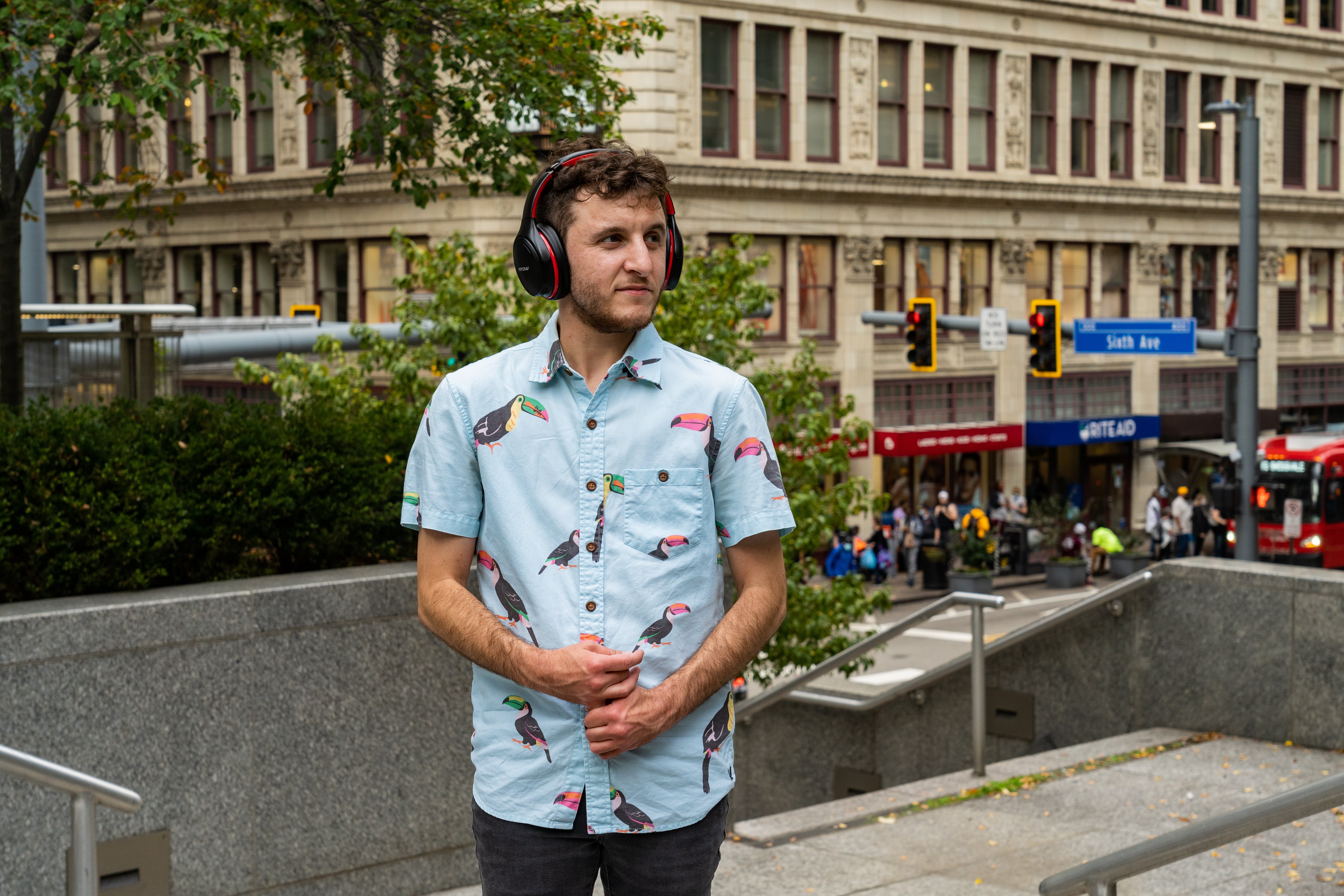 Eli Kurs-Lasky is a white man with curly dark brown hair standing outdoors in a treed plaza with headphones on