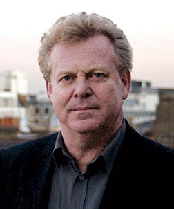 Colin MacCabe is a white man with light red hair, standing on a rooftop with a skyline in the background