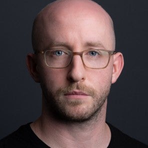 Brett Murphy is a white man wearing brown-framed glasses. His head is shaved and he has a light brown mustache and close beard.
