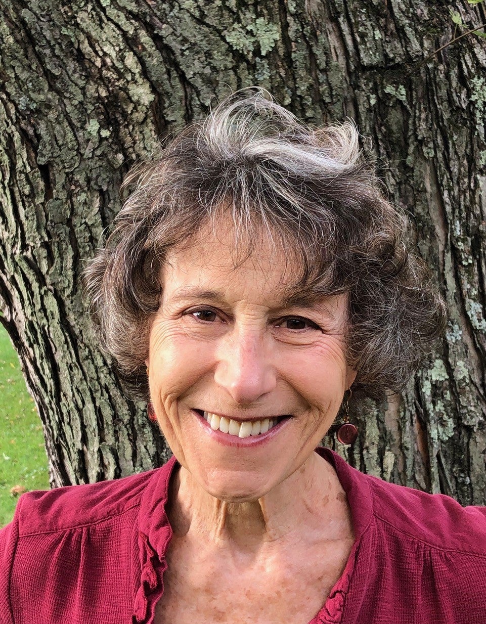 Barbara Edelman, a white woman with chin-length brown hair, standing against a tree trunk.