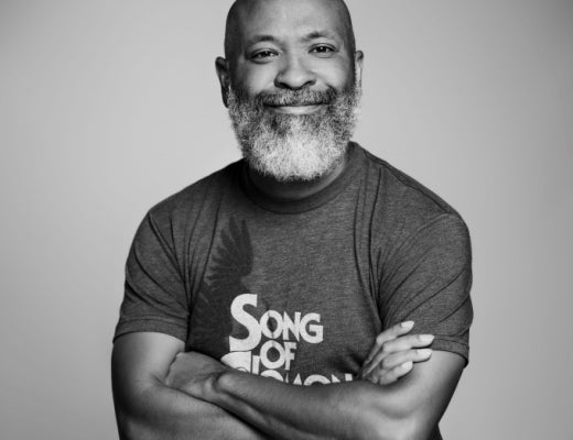 Gary Jackson is a man with medium brown skin and a full gray-white beard/mustache. Black and white photo. Black T-shirt with white lettering (Song of ___), arms folded, against a plain white background