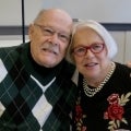 Darlene Kress with her husband Herb. Herb is a light-complexioned older man wearing spectacles and an argyle sweater; to his left, Darlene is light-complexioned, has a white pageboy haircut, and wears red glasses, pears, and a floral print on black background top
