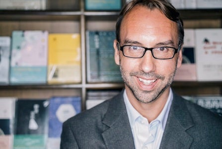 Michael Meyer, a light-skinned man with glasses and facial hair shadow, standing in front of a book display case
