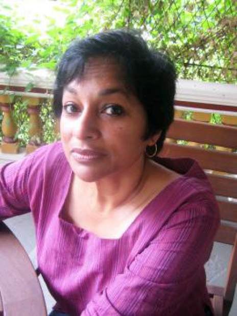 Susan Andrade is brown woman with chin-length black hair sitting at an outdoor patio table, green leaves in background