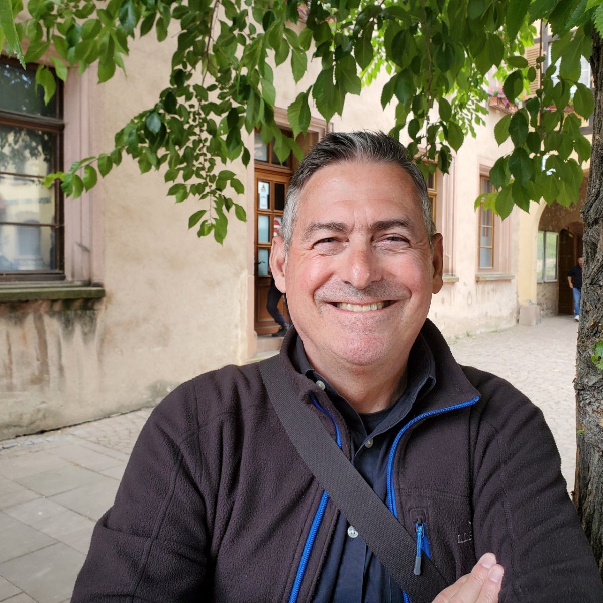 Tom McWhorter, white middle aged mail with salt and pepper hair against a tan stucco buiilding and green leafed tree.